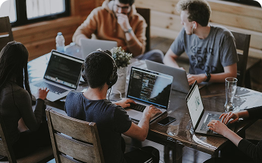 Four young people are sitting around a table with laptops open, working and collaborating in a casual office or meeting space.
