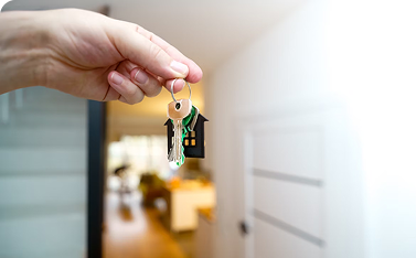 A hand holds up a set of keys on a keychain with a small house-shaped charm, with a blurred, new interior hallway visible in the background.