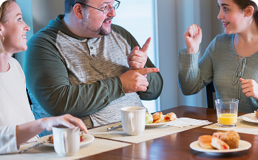 A group of three people sitting at a dining table, smiling and communicating using hand gestures or sign language. Plates of food and drinks are in front of them, creating a warm and friendly atmosphere